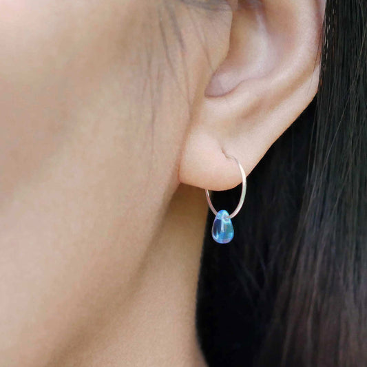 A close up of a sterling silver Czech glass crystal drop in a blue violet teal color on a wire hoop earring, worn outside by a person with dark hair.