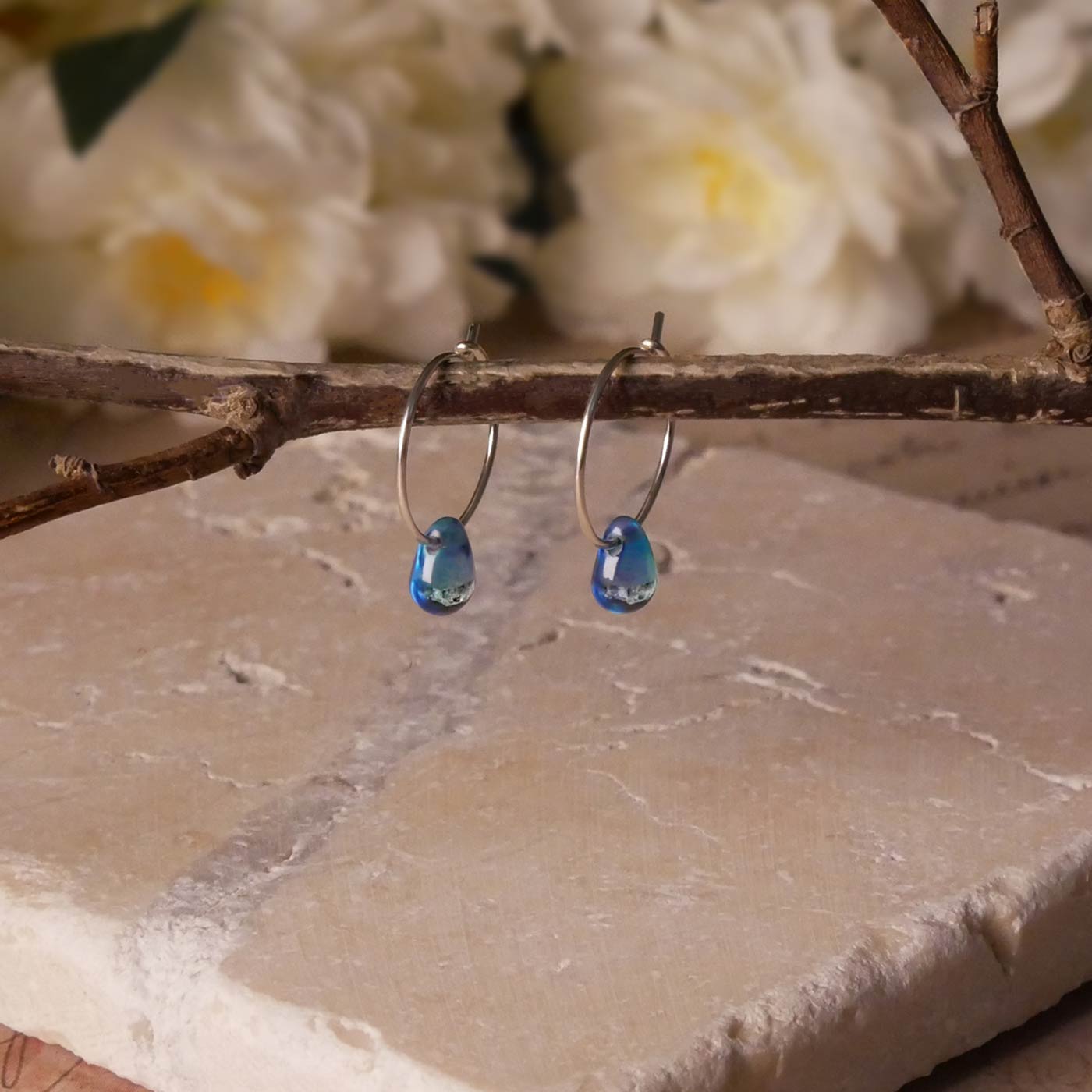 A pair of sterling silver Czech glass crystal blue violet teal colored drop wire hoop earrings, stylized by dangling on a branch over natural beige stone surface, with blurred white flowers in the background.