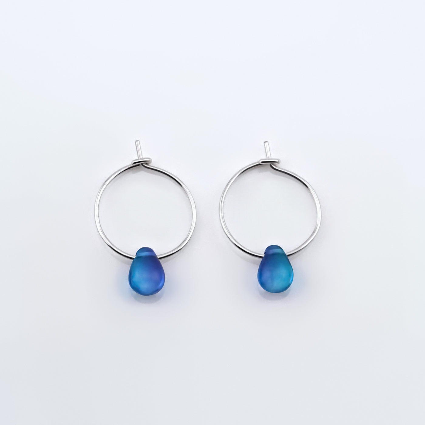 Top view of a pair of sterling silver Czech glass crystal blue violet teal colored drop wire hoop earrings, laying flat on a plain white background.