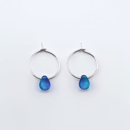 Top view of a pair of sterling silver Czech glass crystal blue violet teal colored drop wire hoop earrings, laying flat on a plain white background.