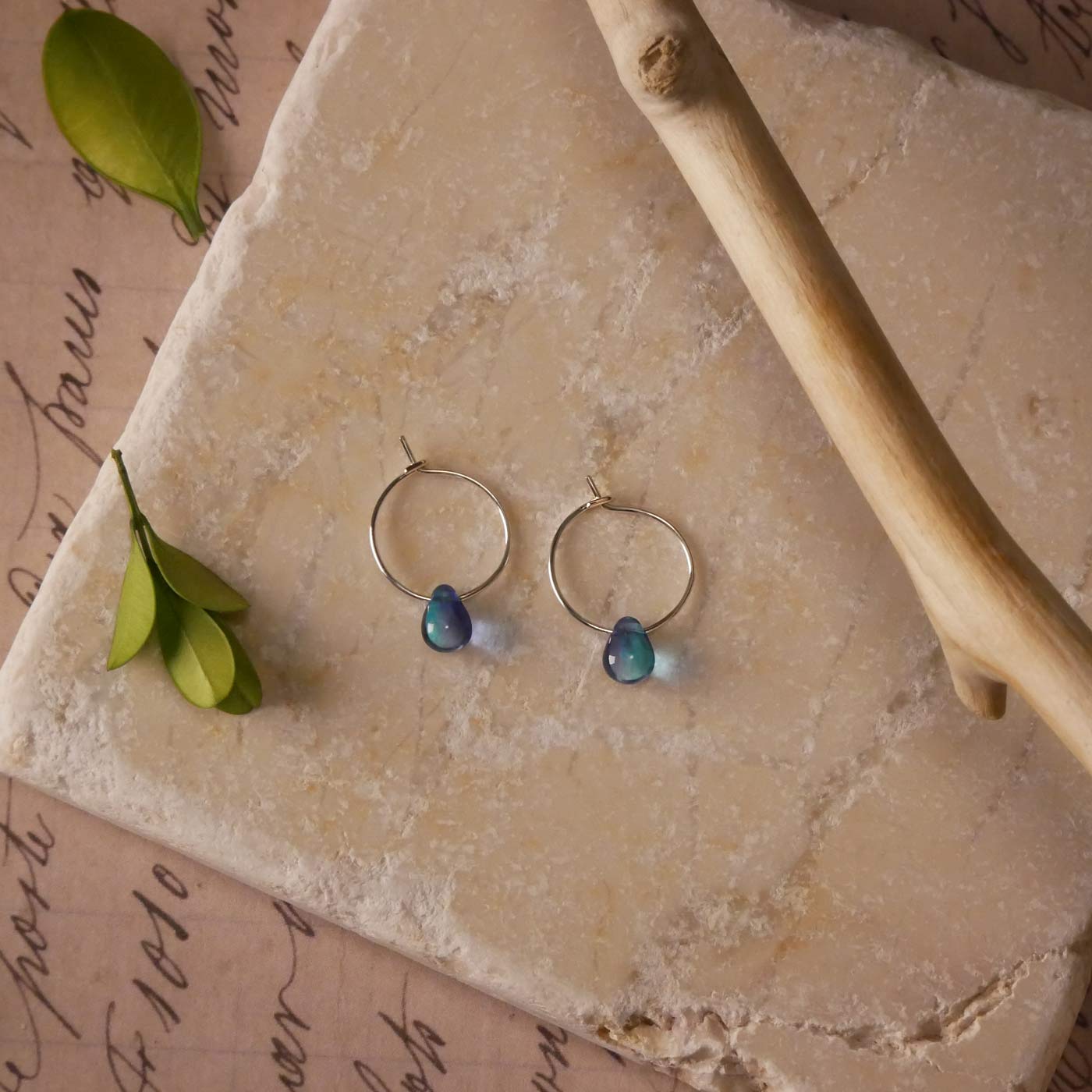 Top view with a pair of sterling silver Czech glass crystal, blue violet colored drop wire hoop earrings. It is stylized by laying flat on a natural stone surface, over vintage paper with cursive writing, and surrounded by green leaves with a branch in the foreground.