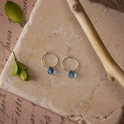 Top view with a pair of sterling silver Czech glass crystal, blue violet colored drop wire hoop earrings. It is stylized by laying flat on a natural stone surface, over vintage paper with cursive writing, and surrounded by green leaves with a branch in the foreground.