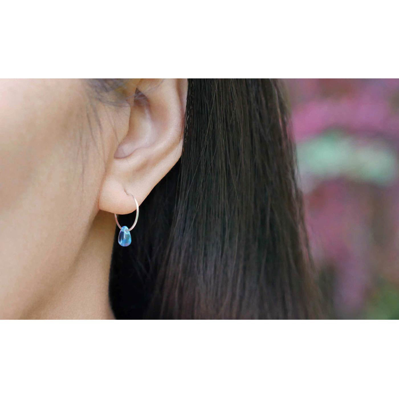 A close up of a sterling silver Czech glass crystal blue violet colored drop wire hoop earring, worn outside by a person with dark hair. The background is blurred with plants in the background.