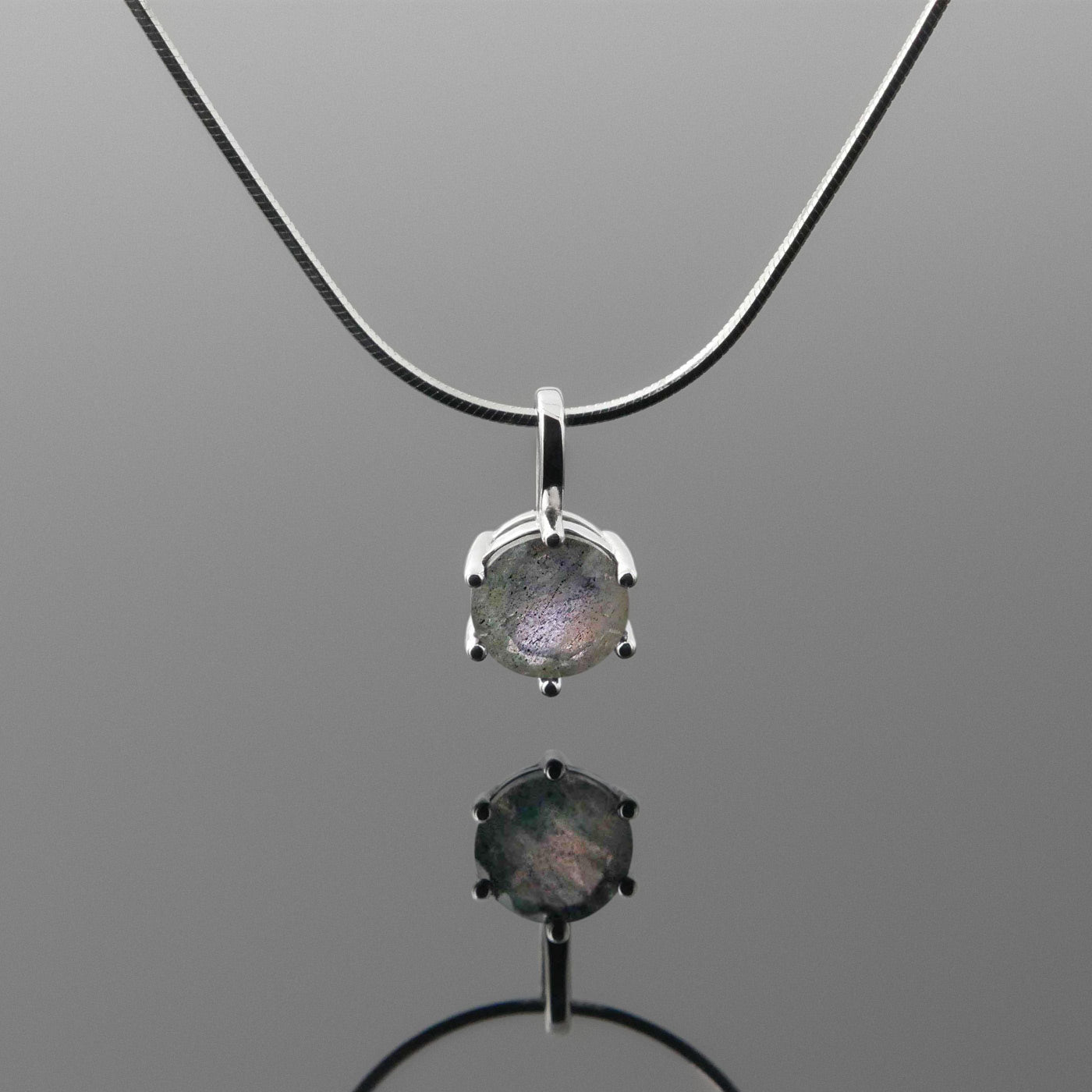 Front view of a dainty, round high quality Labradorite prong set pendant sterling silver necklace hanging, with a plain reflective gray background.
