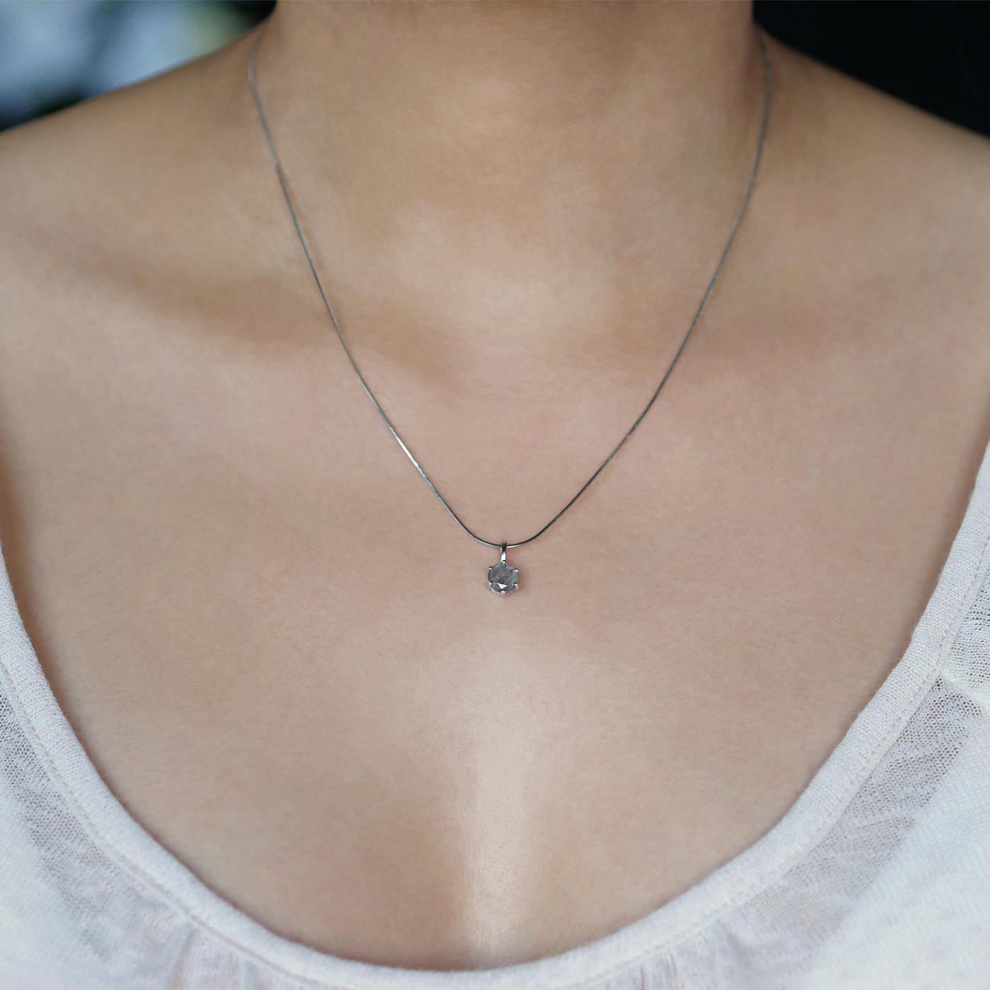 Front view of a dainty, round high quality Labradorite prong set sterling silver pendant necklace worn by a person with a beige top, against a blurred dark background with white flowers.