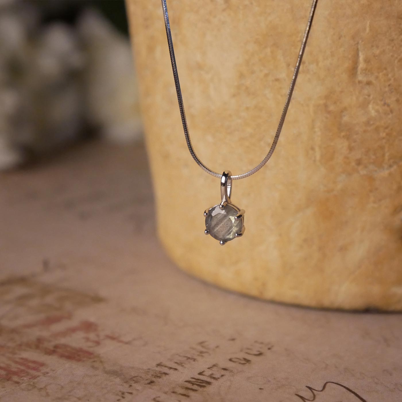 Front view of a dainty, round high quality Labradorite prong set, sterling silver pendant necklace. It is stylized by hanging from a textured light brown pot, over vintage paper with old writing style on it, with white flowers blurred in background.