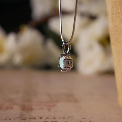 Side view of a dainty, round high quality Labradorite prong set, sterling silver pendant necklace. It is stylized by hanging from a textured light brown pot, over vintage paper with old writing style on it, with white flowers blurred in background.