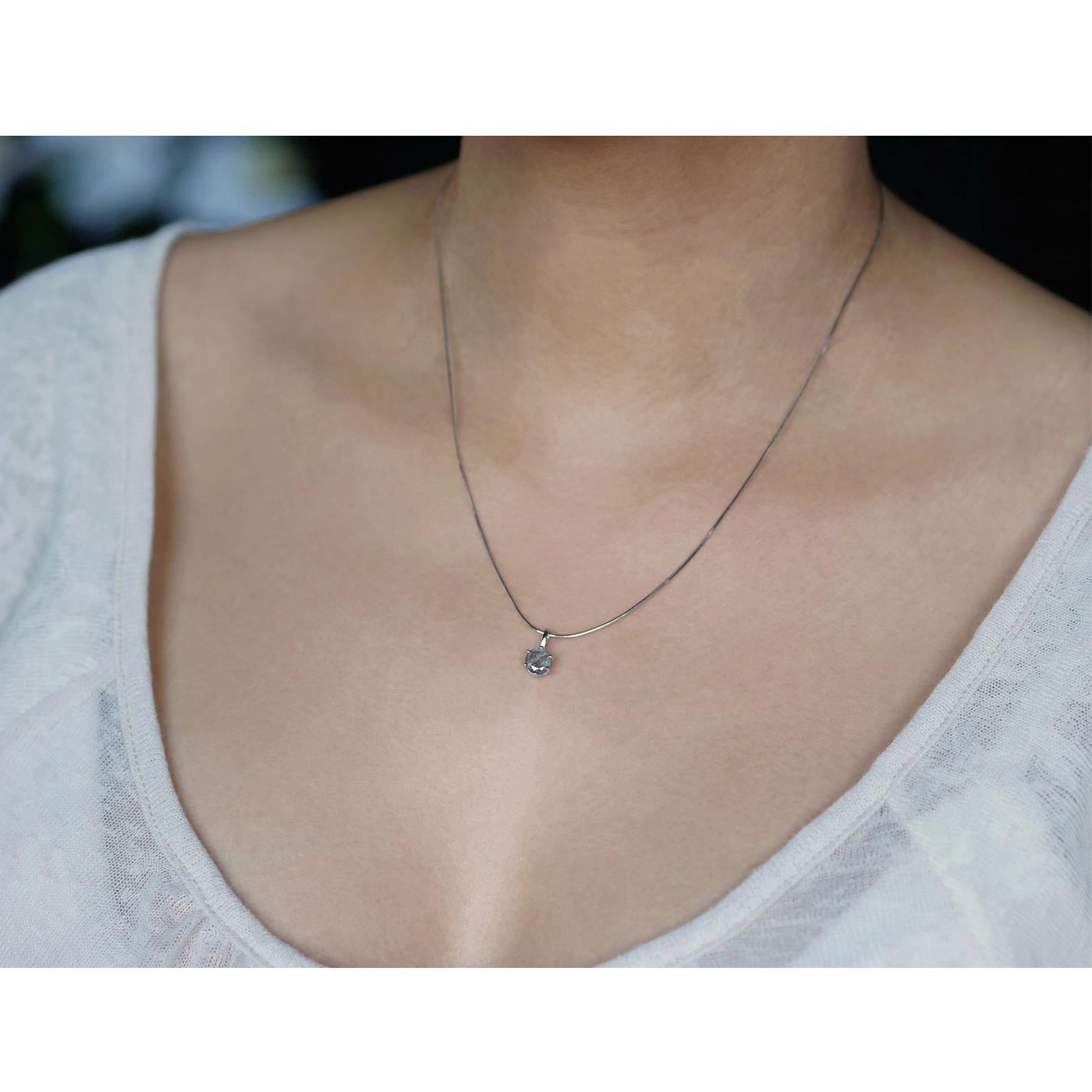 Front view of a dainty, round high quality Labradorite prong set sterling silver pendant necklace worn by a person with a beige top, against a blurred dark background with white flowers.