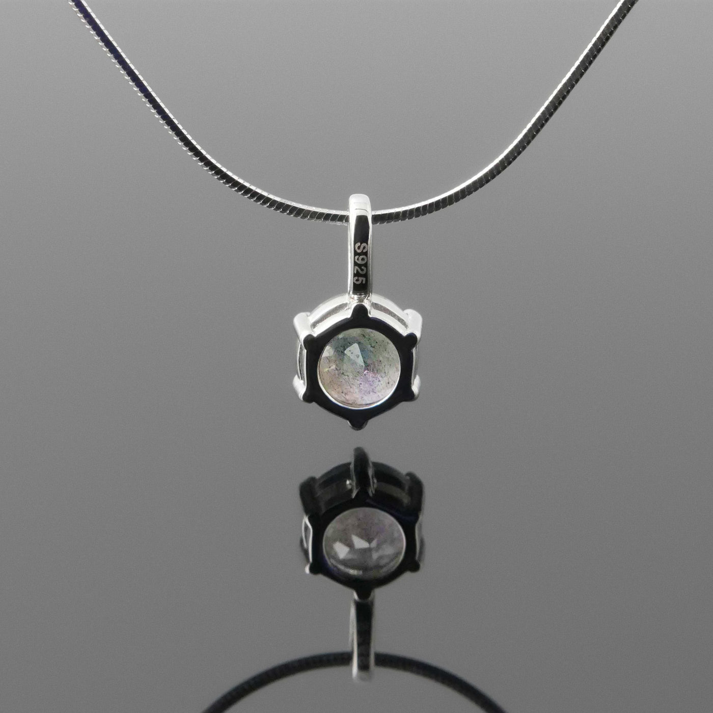 Back view of a dainty, round high quality Labradorite prong set pendant sterling silver necklace hanging, with a plain reflective gray background.