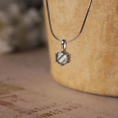 Front view of a dainty, round high quality Labradorite prong set, sterling silver pendant necklace. It is stylized by hanging from a textured light brown pot, over vintage paper with old writing style on it, with white flowers blurred in background.