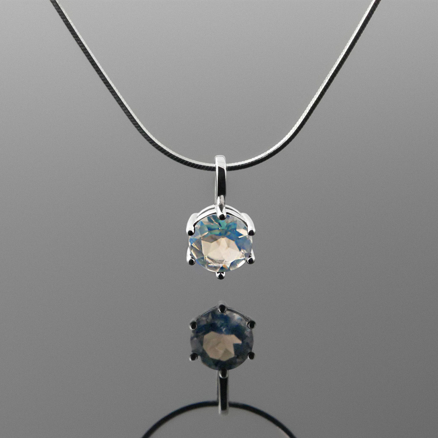 Front view of a dainty, round high quality eye clean faceted AAA Rainbow Blue Moonstone prong set pendant sterling silver necklace hanging, with a plain reflective gray background.
