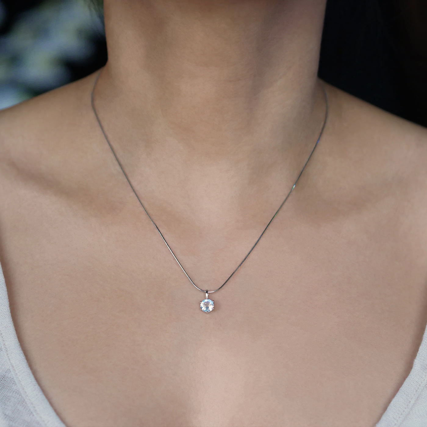 Front view of a dainty, round high quality eye clean faceted AAA Rainbow Moonstone prong set  sterling silver pendant necklace worn by a person with a beige top, against a blurred dark background with white flowers.