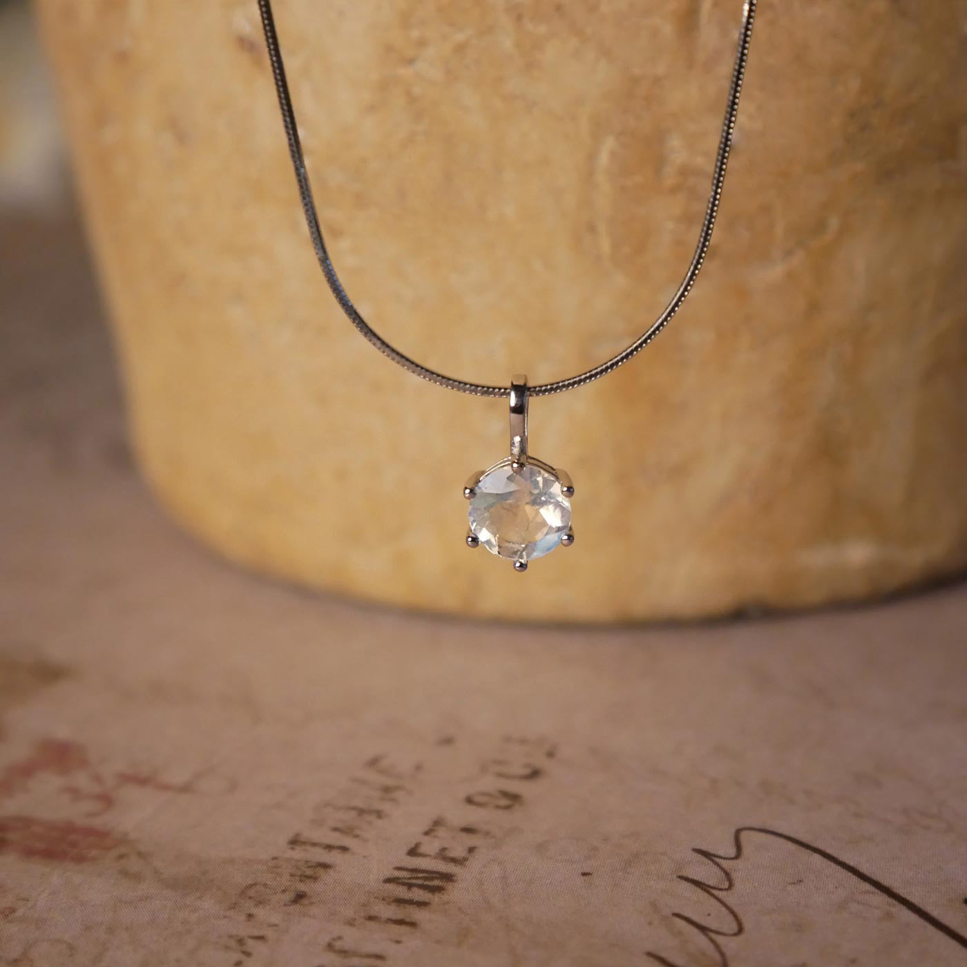 Front view of a dainty, round high quality eye clean faceted AAA Rainbow Blue Moonstone prong set sterling silver pendant necklace. It is stylized by hanging from a textured light brown pot, over vintage paper with old writing style on it.
