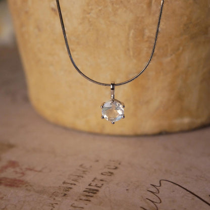 Front view of a dainty, round high quality eye clean faceted AAA Rainbow Blue Moonstone prong set sterling silver pendant necklace. It is stylized by hanging from a textured light brown pot, over vintage paper with old writing style on it.