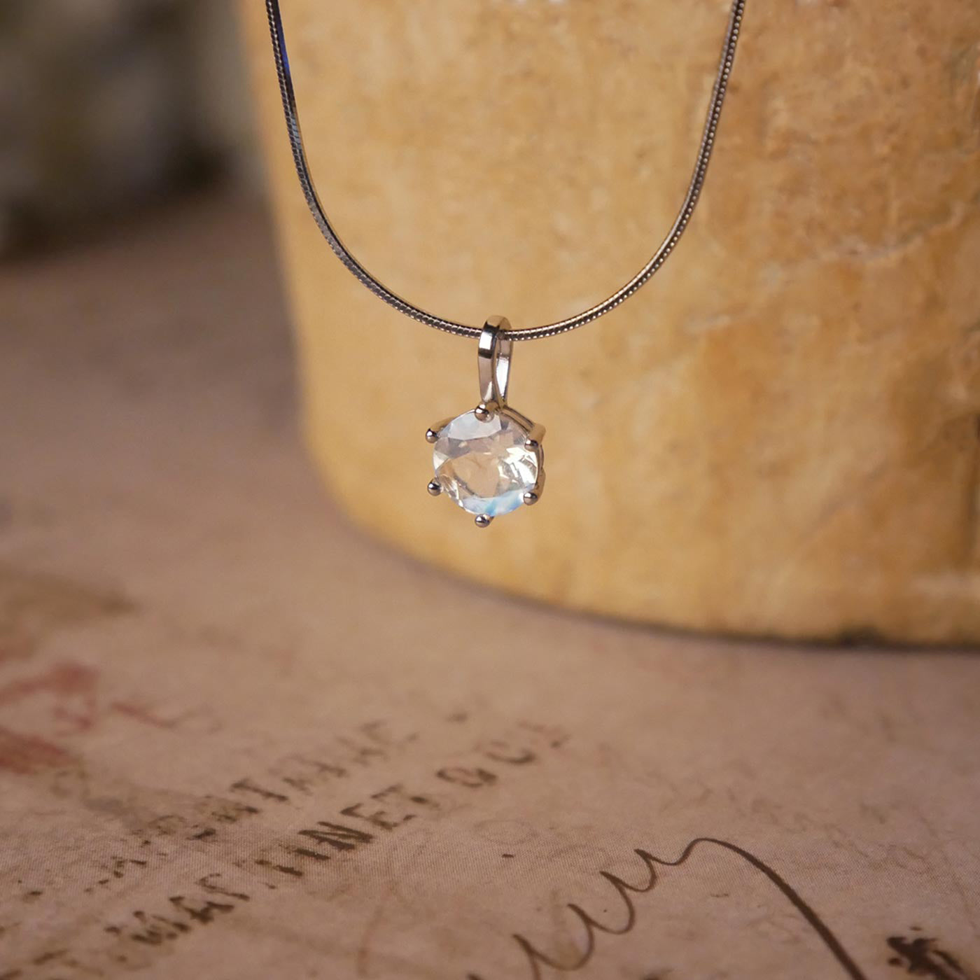 Front view of a dainty, round high quality eye clean faceted AAA Rainbow Blue Moonstone prong set sterling silver pendant necklace. It is stylized by hanging from a textured light brown pot, over vintage paper with old writing style on it.