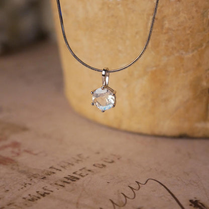 Front view of a dainty, round high quality eye clean faceted AAA Rainbow Blue Moonstone prong set sterling silver pendant necklace. It is stylized by hanging from a textured light brown pot, over vintage paper with old writing style on it.
