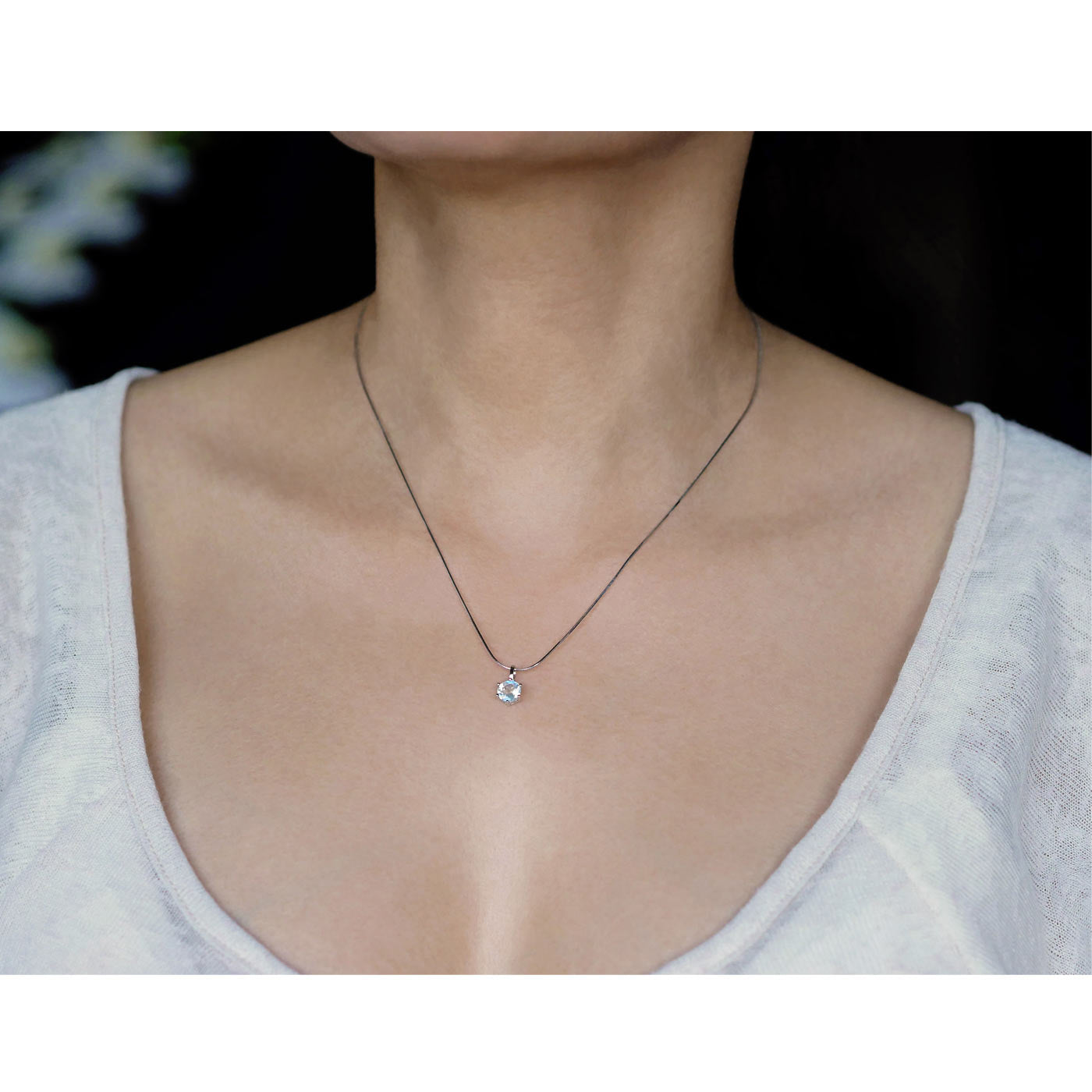 Front view of a dainty, round high quality eye clean faceted AAA Rainbow Moonstone prong set sterling silver pendant necklace worn by a person with a beige top, against a blurred dark background with white flowers.