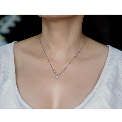 Front view of a dainty, round high quality eye clean faceted AAA Rainbow Moonstone prong set sterling silver pendant necklace worn by a person with a beige top, against a blurred dark background with white flowers.