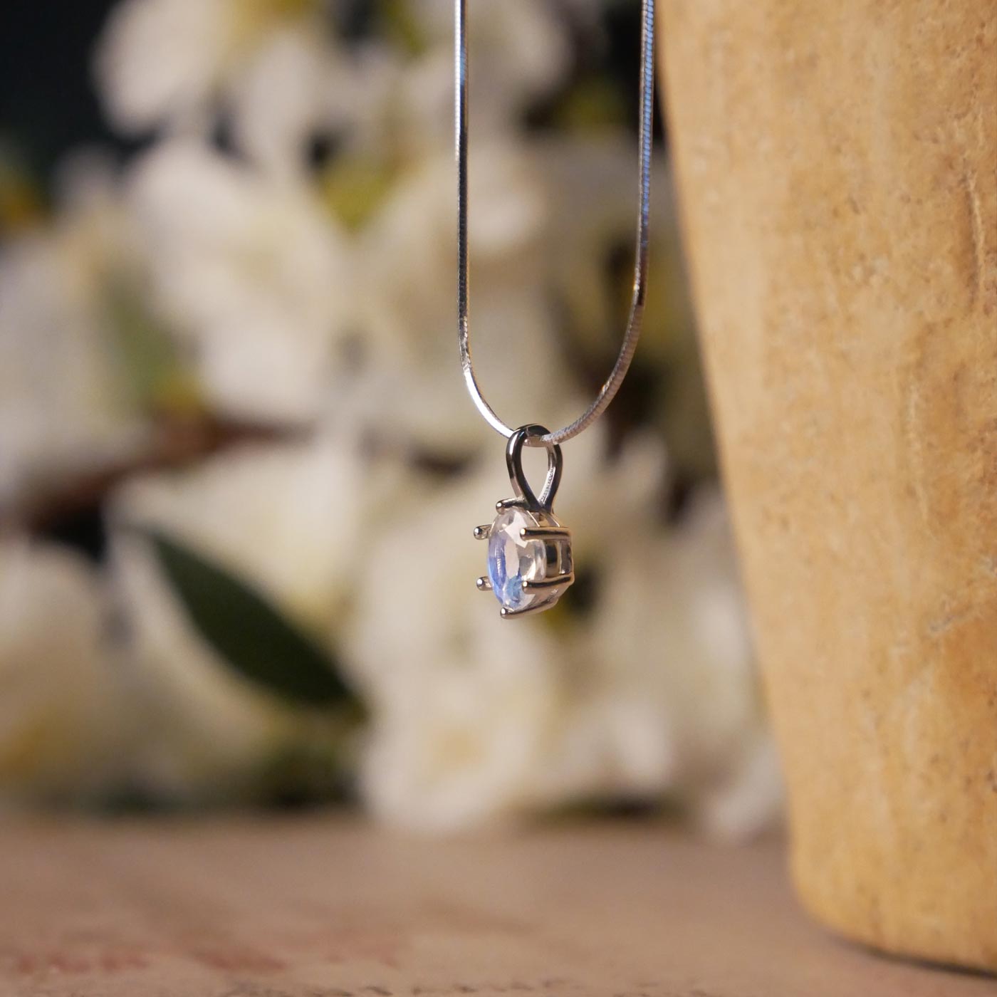 Side view of a dainty, round high quality eye clean faceted AAA Rainbow Blue Moonstone prong set sterling silver pendant necklace. It is stylized by hanging from a textured light brown pot, over vintage paper with old writing style on it, with white flowers blurred in background.