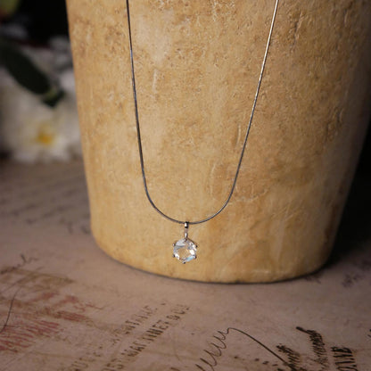 Front view of a dainty, round high quality eye clean faceted AAA Rainbow Blue Moonstone prong set sterling silver pendant necklace. It is stylized by hanging from a textured light brown pot, over vintage paper with old writing style on it, with white flowers blurred in background.