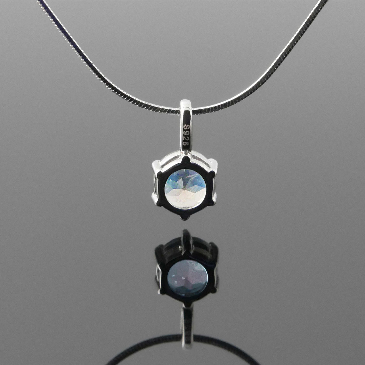 Back view of a dainty, round high quality eye clean faceted AAA Rainbow Blue Moonstone prong set pendant sterling silver necklace hanging, with a plain reflective gray background.
