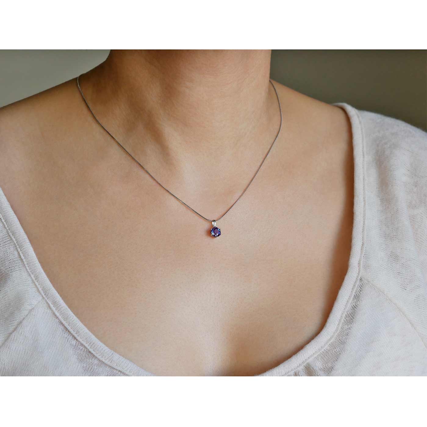3/4 view of a dainty, round high quality eye clean faceted AAA Chrysoberyl lab grown genuine authentic Alexandrite prong set pendant sterling silver necklace worn by a person with a beige top, against a plain beige gray background.