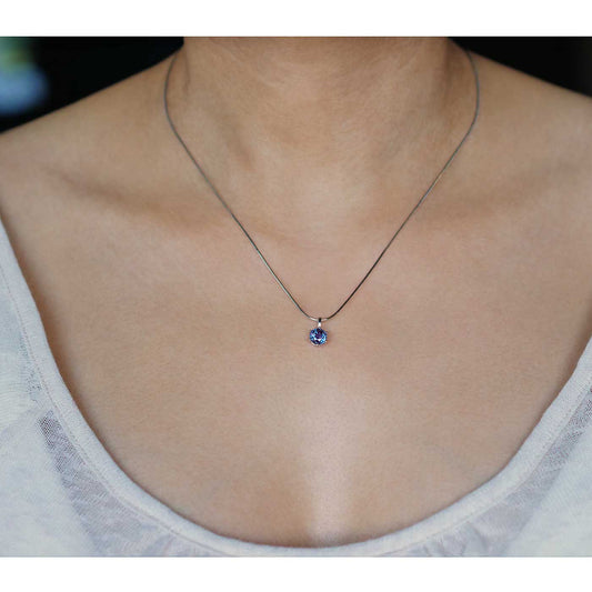 Front view of a dainty, round high quality eye clean faceted AAA Chrysoberyl lab grown genuine authentic Alexandrite prong set pendant sterling silver necklace worn by a person with a beige top, against a blurred dark background with white flowers.
