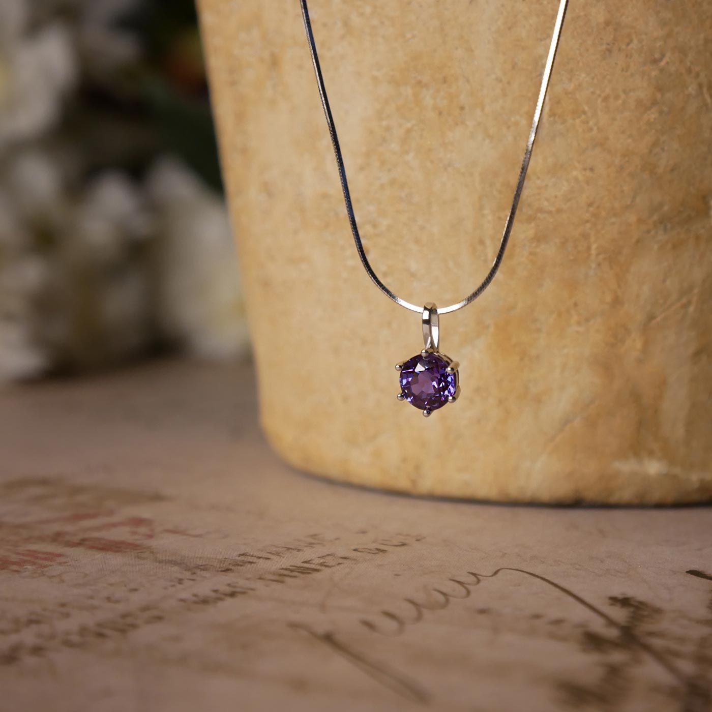 Front view of dainty, round high quality eye clean faceted AAA Chrysoberyl lab grown genuine authentic Alexandrite prong set pendant sterling silver necklace. It is stylized by hanging from a textured light brown pot, over vintage paper, with old writing style on it and white flowers blurred in the background.