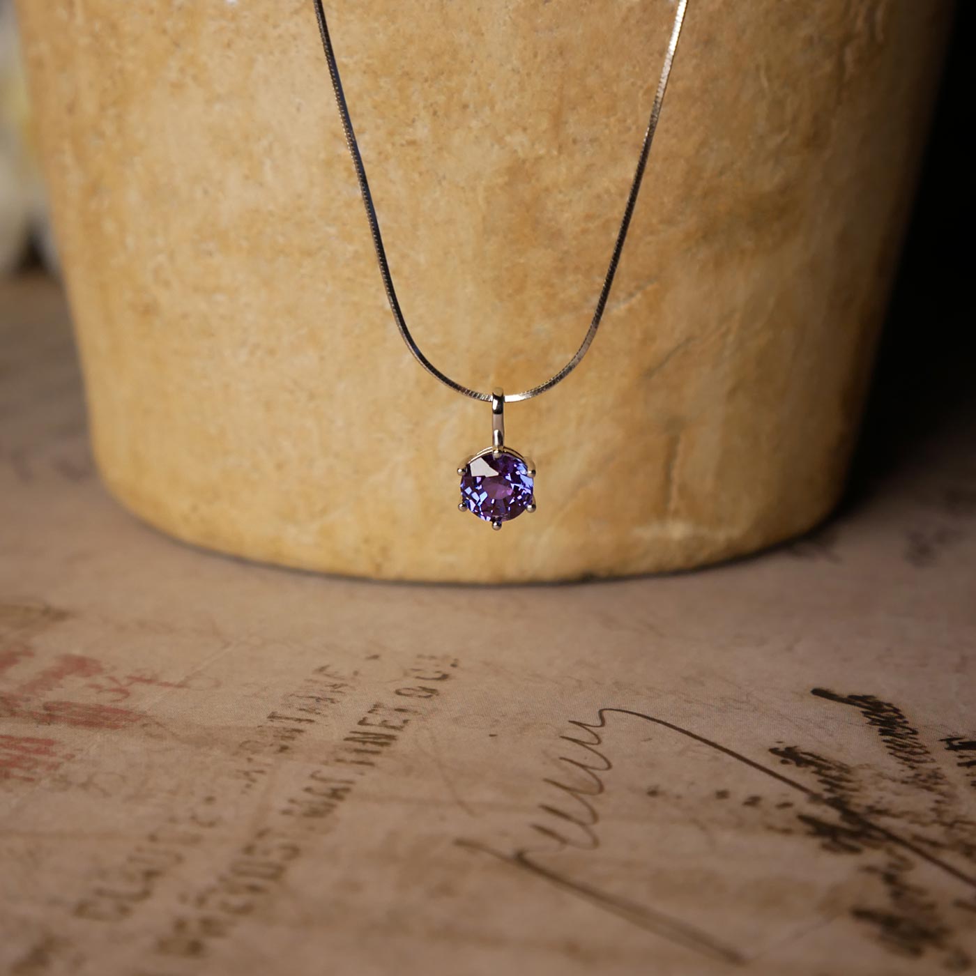 Front view of dainty, round high quality eye clean faceted AAA Chrysoberyl lab grown genuine authentic Alexandrite prong set pendant sterling silver necklace. It is stylized by hanging from a textured light brown pot, over vintage paper, with old writing style on it and white flowers blurred in the background.