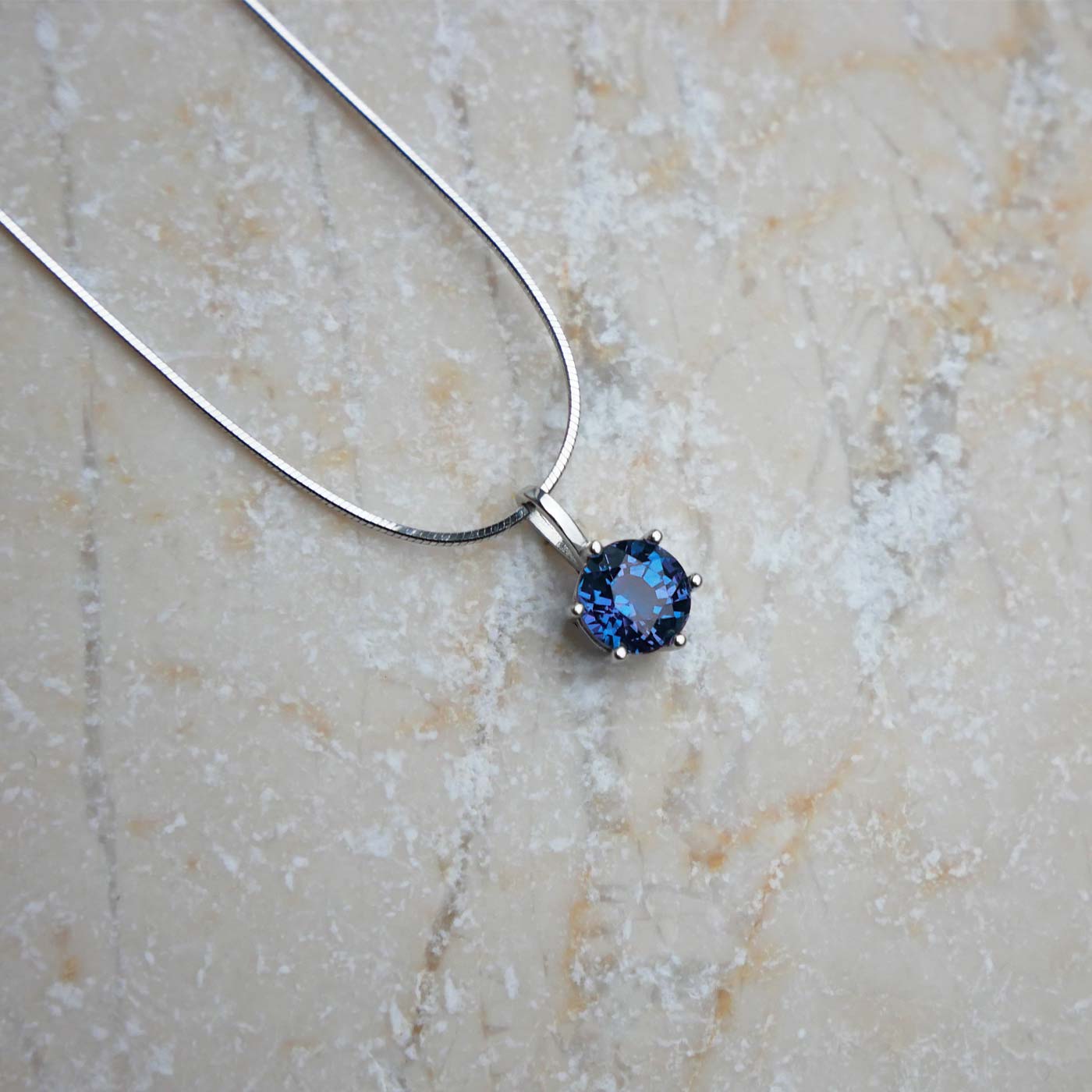 Front view of dainty, round high quality eye clean faceted AAA Chrysoberyl lab grown genuine authentic Alexandrite prong set pendant sterling silver necklace laying flat on a beige textured stone surface.