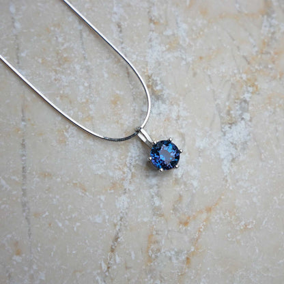 Front view of dainty, round high quality eye clean faceted AAA Chrysoberyl lab grown genuine authentic Alexandrite prong set pendant sterling silver necklace laying flat on a beige textured stone surface.
