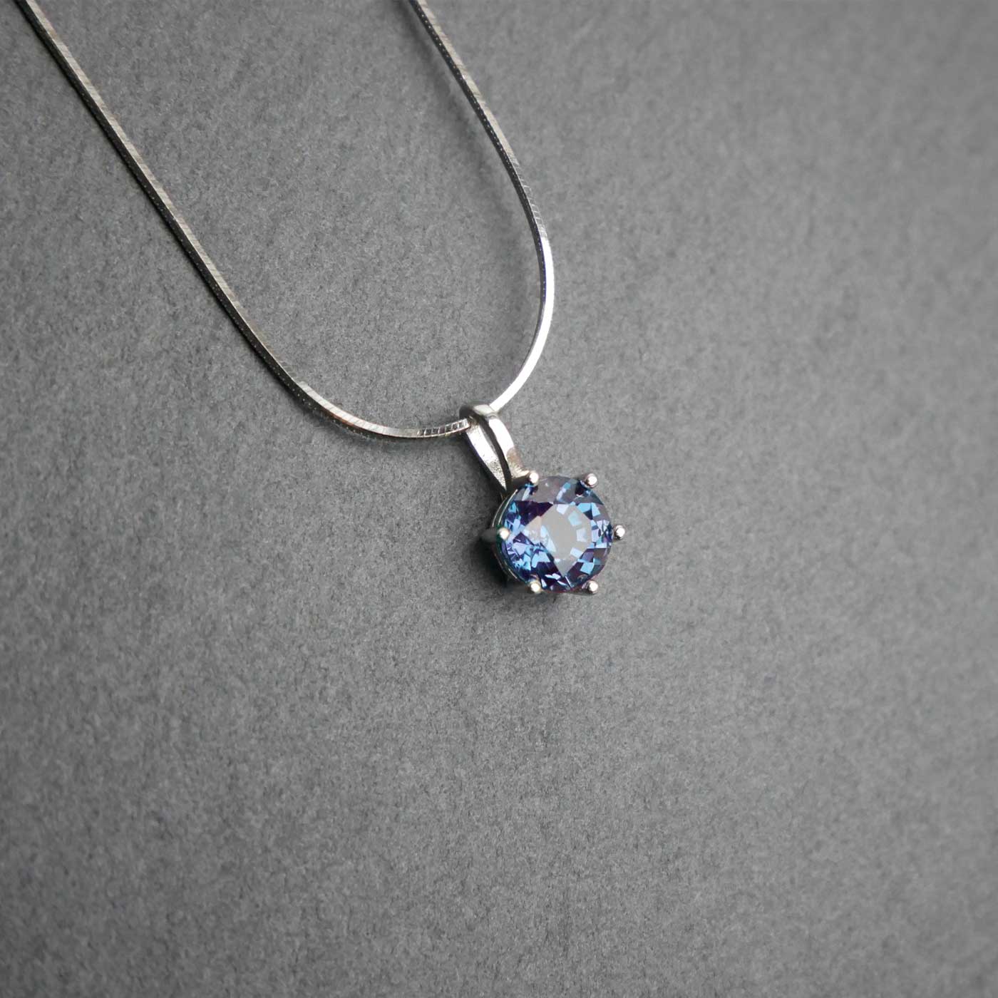 Front view of dainty, round high quality eye clean faceted AAA Chrysoberyl lab grown genuine authentic Alexandrite prong set pendant sterling silver necklace laying flat on a dark gray slate textured stone surface.
