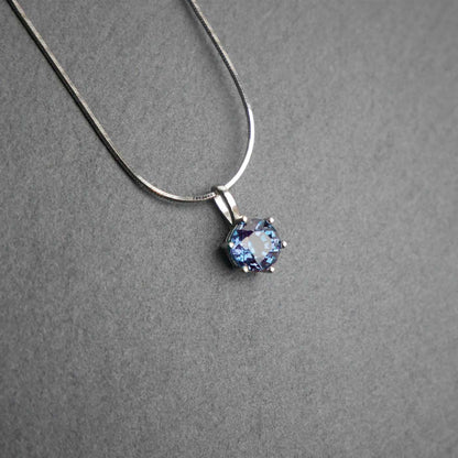 Front view of dainty, round high quality eye clean faceted AAA Chrysoberyl lab grown genuine authentic Alexandrite prong set pendant sterling silver necklace laying flat on a dark gray slate textured stone surface.