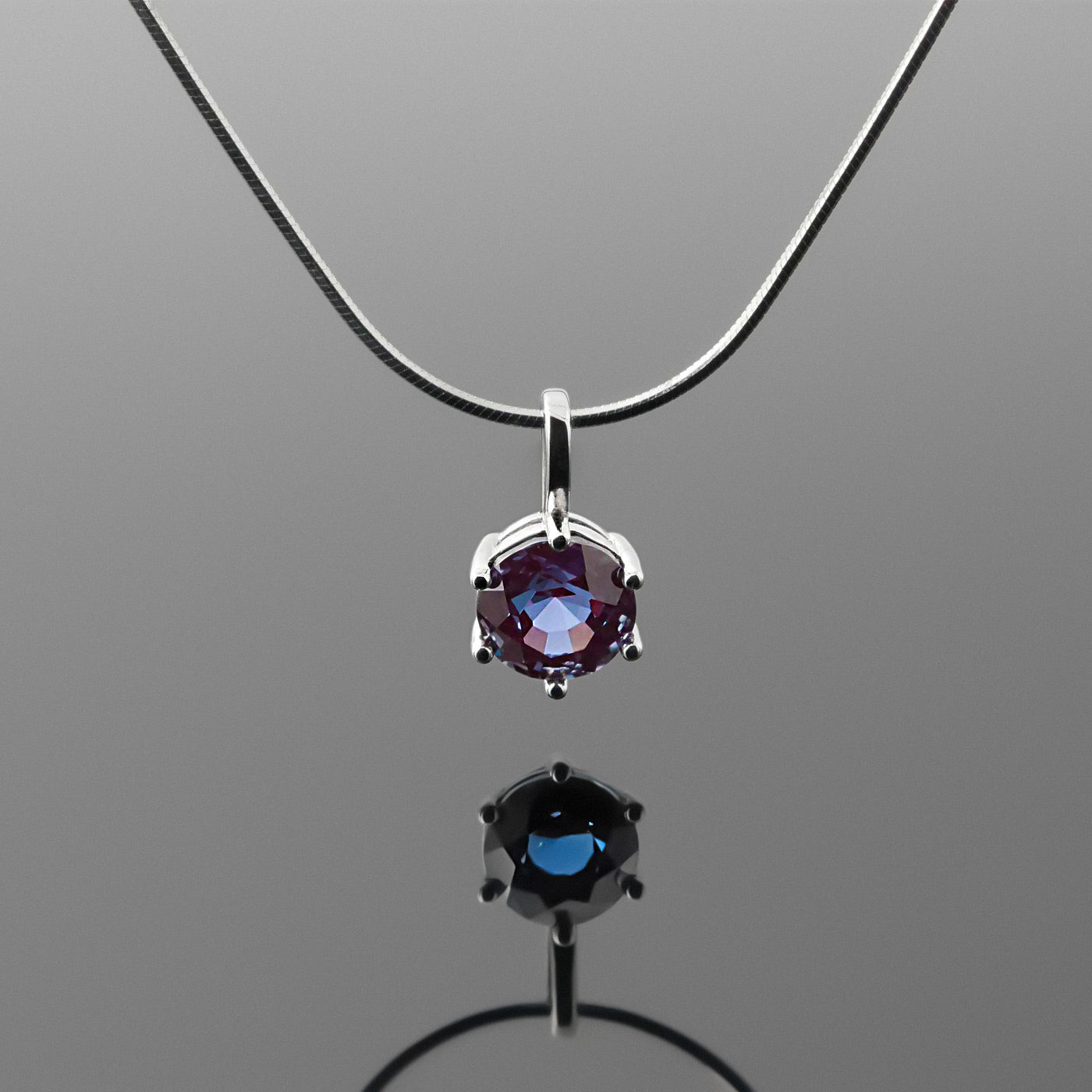 Front view of a dainty, round high quality eye clean faceted AAA Chrysoberyl lab grown genuine authentic Alexandrite prong set pendant sterling silver necklace hanging, with a plain reflective gray background.